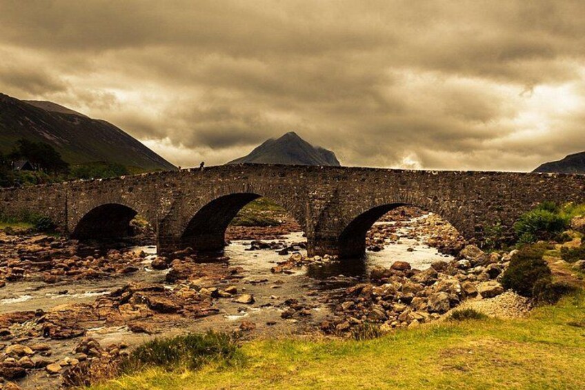 A stone bridge with three arches over a rocky riverbed, surrounded by grassy terrain and mountains in the background. 2