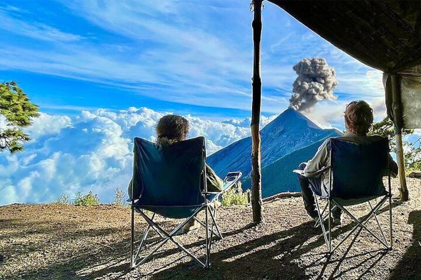 Deux personnes sont assises sur des chaises pliantes, et devant elles se dresse une montagne sur laquelle s’élève une colonne de fumée. 2