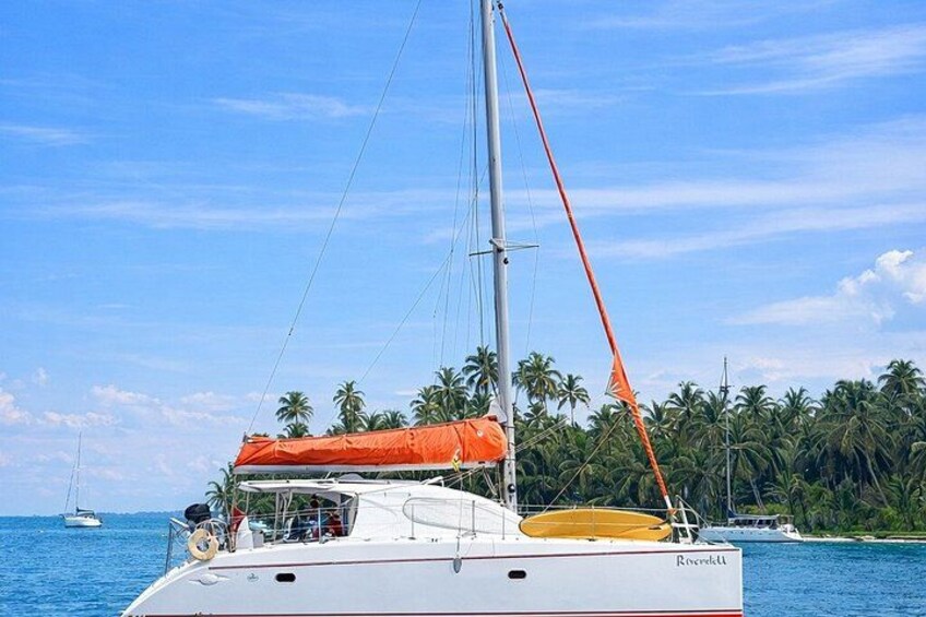 A sailboat named 'Rosella' with a red canopy and a yellow lifebuoy on the deck, sailing near a beach with palm trees and a clear blue sky. 2