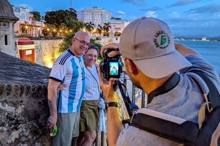 A person taking a photo of two people standing by a waterfront with buildings in the background. 5