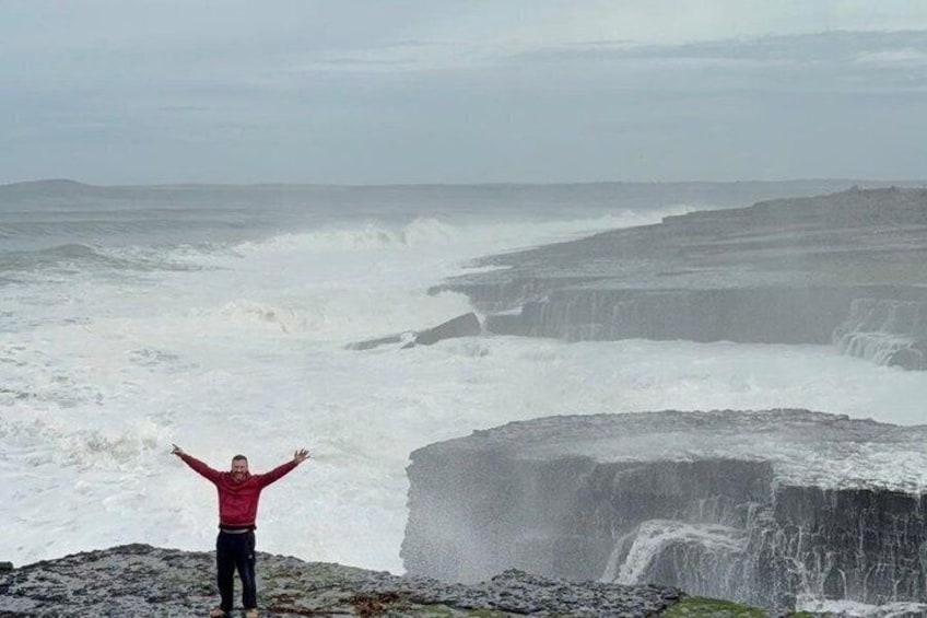 A person standing on a rocky outcrop with arms raised, overlooking a coastal area with waves crashing against a cliff. 3