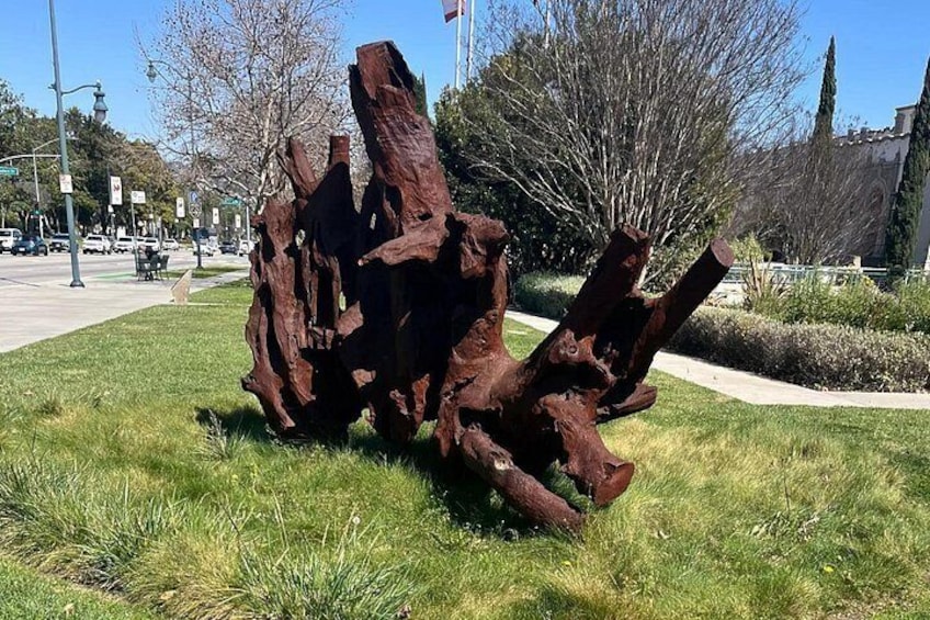A large, rust-colored sculpture of tree trunks in a grassy area with a sidewalk and buildings in the background. 5