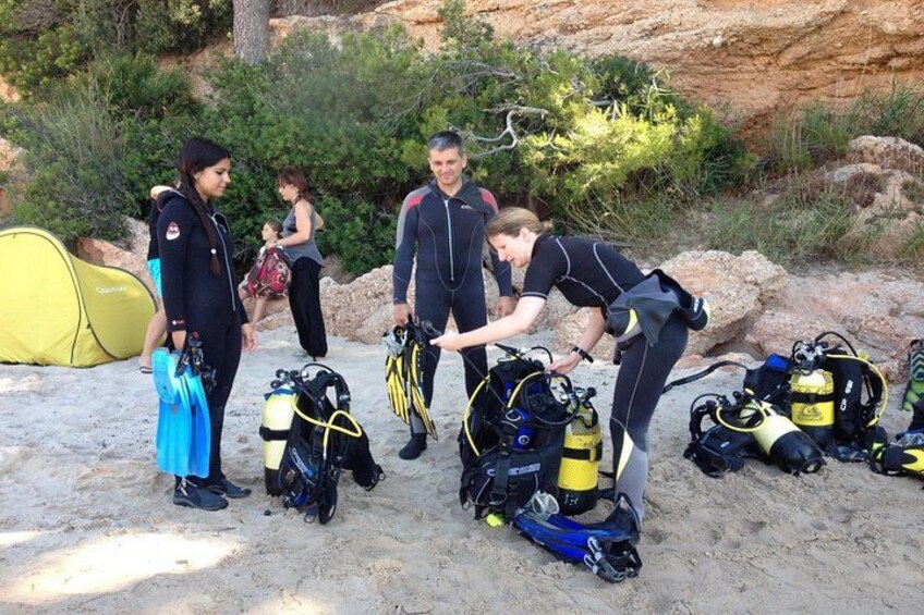 A group of people preparing scuba diving equipment on a sandy beach with a tent and rocky terrain in the background. 3