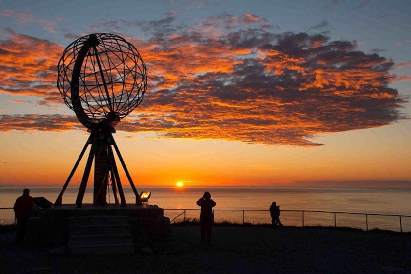 En solnedgång med en sfärisk skulptur på ett stativ, silhuettliknande figurer på stranden och havet. 4