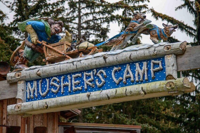 Et træskilt med teksten 'MUSHER'S CAMP' og dekorative figurer af bjørne og en hund. 1