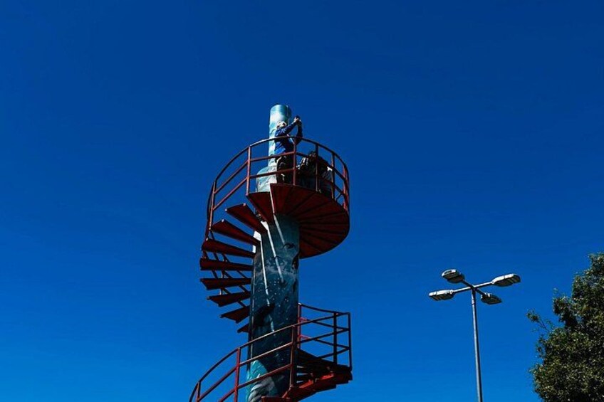 A tall tower with a spiral staircase and a cylindrical structure at the top against a clear blue sky. 5