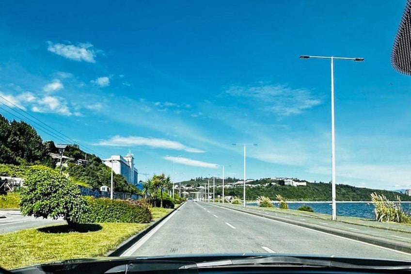 A road with a clear blue sky, greenery, a body of water, and distant buildings. 4