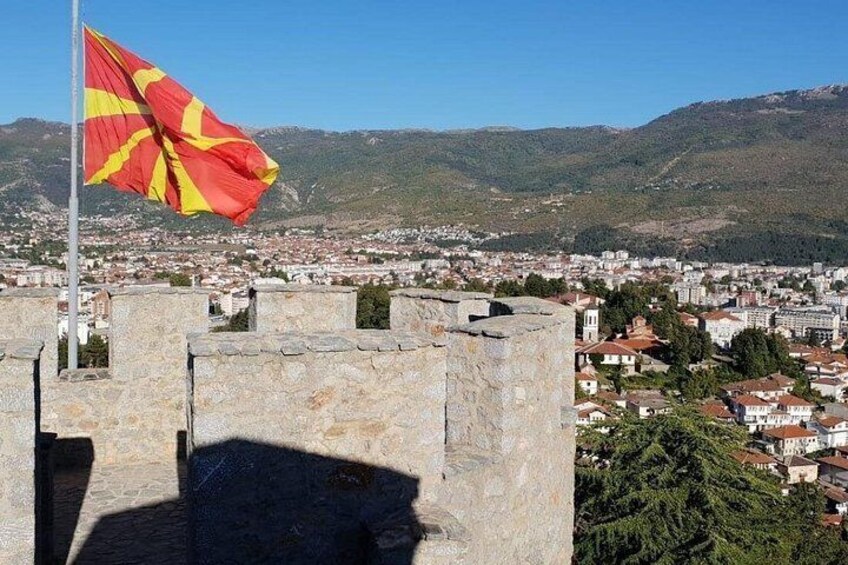 A flag on a flagpole, a stone wall, and a cityscape with buildings and mountains in the background. 1