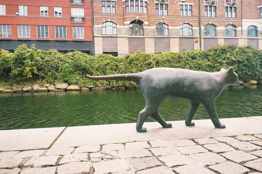 A statue of a cat walking along a paved path beside a body of water, with buildings in the background. 2