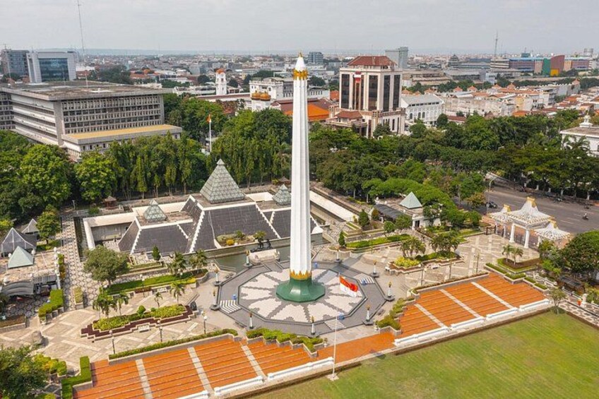 A tall monument with a green base, surrounded by a plaza with patterned paving, trees, and buildings in the background. 2