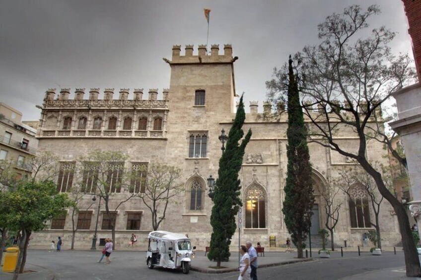 A historic building with a tower, large windows, and a flag. There are trees and a street with people and a vehicle. 1