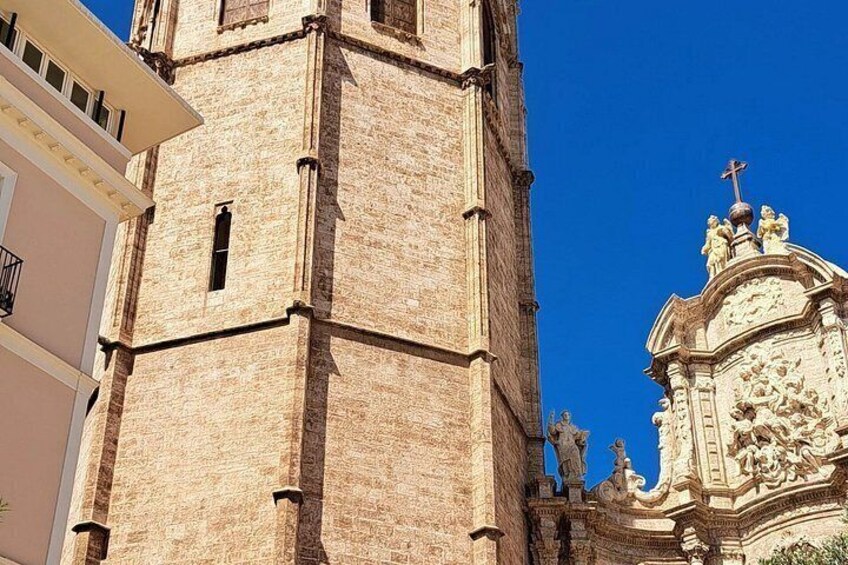 A tall brick tower with a cross on top, adjacent to a building with a balcony. 5