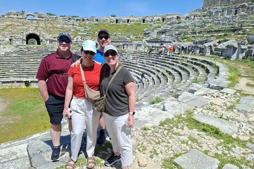 A group of people standing in front of an ancient amphitheater with stone seating and a partially visible stage. 2