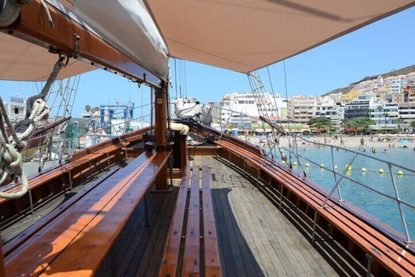 A wooden deck of a sailing ship with ropes and rigging, overlooking a coastal town with buildings and a beach. 3