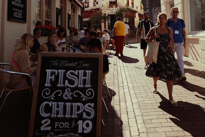 En gade med mennesker, der spiser ved udendørs borde, et skilt, der reklamerer for fish and chips, og en bygning med et skilt for ales. 2