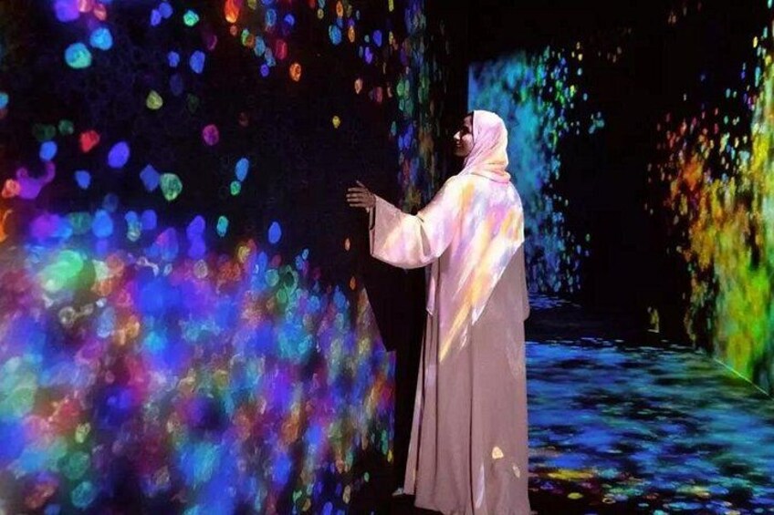 Une personne vêtue d’un costume traditionnel se tient dans une pièce dont les murs sont illuminés par des projections de lumière colorée. 2