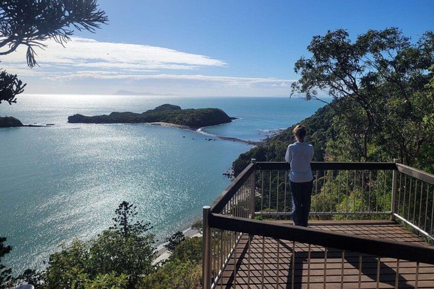 A person standing on a wooden deck overlooking a coastal landscape with clear blue water and distant islands. 2