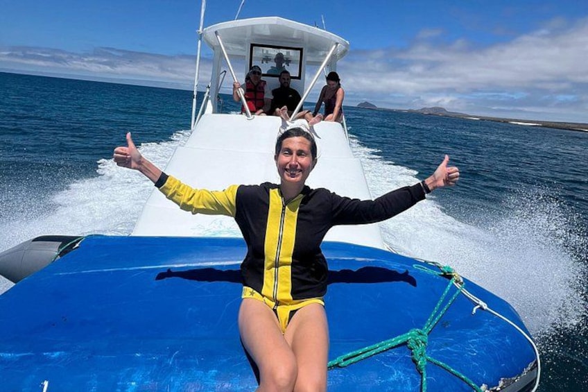 A person in a yellow and black wetsuit sitting on a blue boat with arms outstretched, a rope tied to the boat, and another person in the background. 1