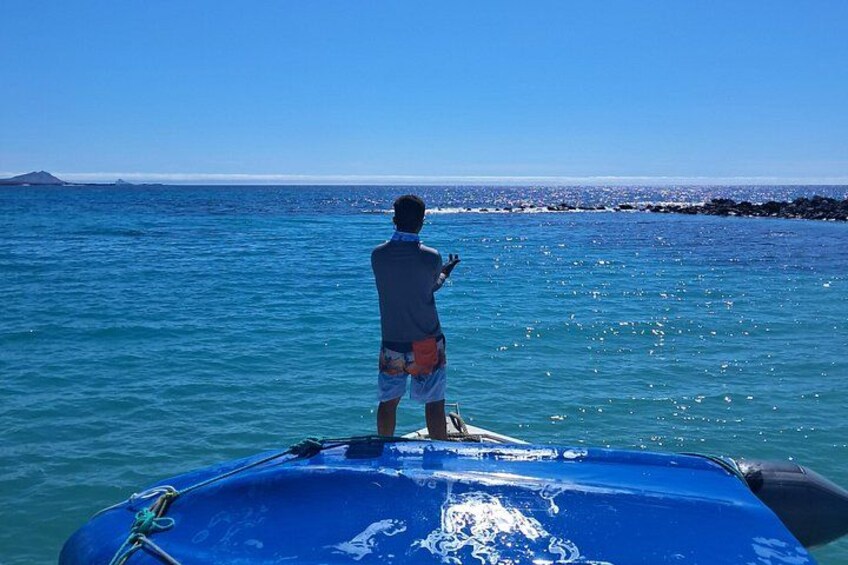 A person standing on a blue boat in clear blue water, with a rocky shoreline and a distant landmass in the background. 4