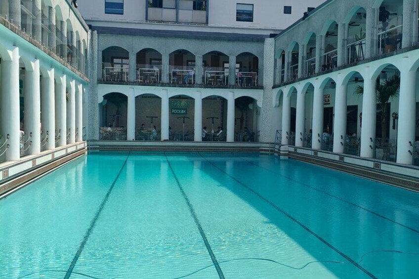 Ein großes Hallenbad mit klarem, blauem Wasser, umgeben von weißen Säulen und Balkonen. 4