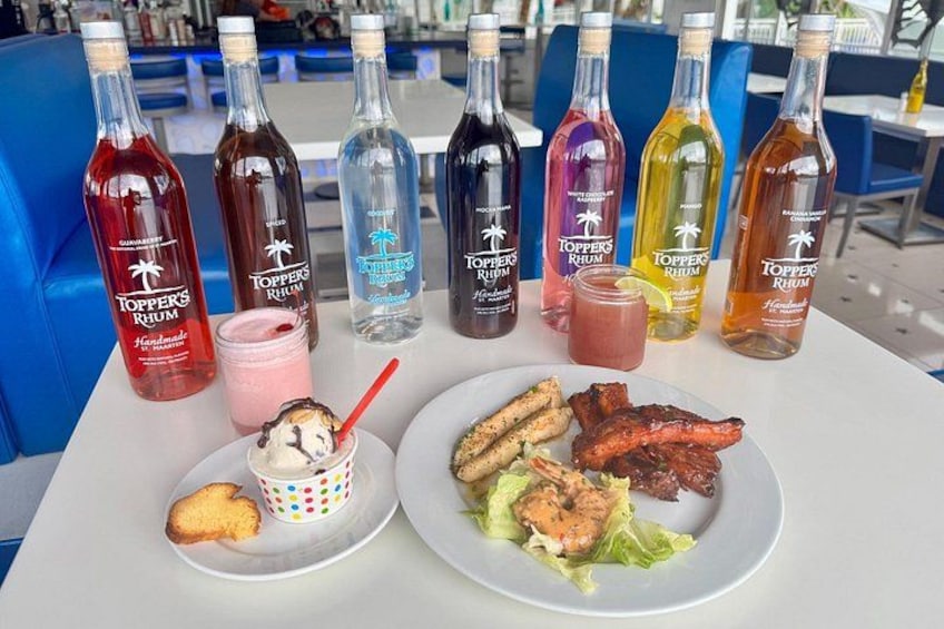 A table with various bottles of Toppers Rum, a dessert with a red spoon, and plates of food including grilled meat and vegetables. 1