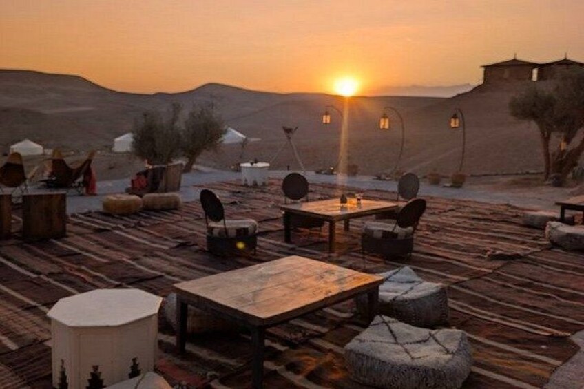 A desert campsite with wooden tables and chairs, tents, and a sunset in the background. 2