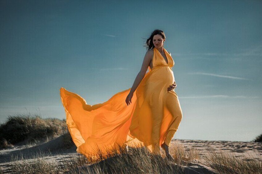 A pregnant woman in a flowing yellow dress walking on a sandy beach with dunes in the background. 2