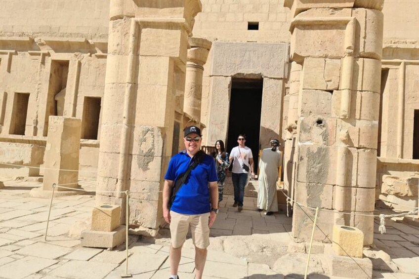 A man in a blue shirt and beige shorts standing in front of ancient stone architecture with columns and a doorway. 2