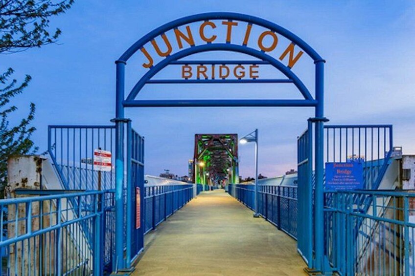 Une arche métallique bleue, portant l’inscription « JUNCTION BRIDGE », avec une passerelle en béton menant à une structure située au loin. 2