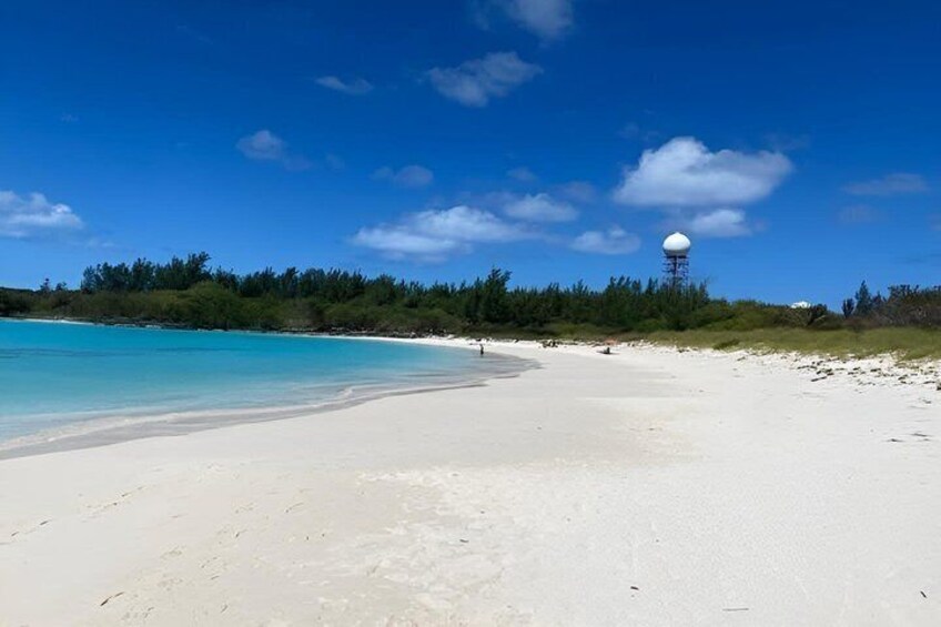 A beach with clear turquoise water, white sand, and a water tower in the distance. 2