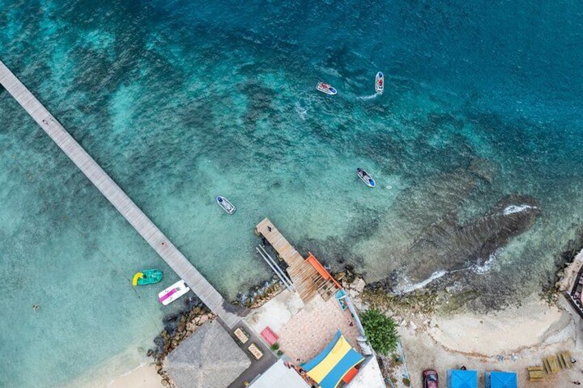 Luchtbeeld van een kustgebied met een pier die zich uitstrekt over turkoois water, verschillende boten en strandparasols. 1