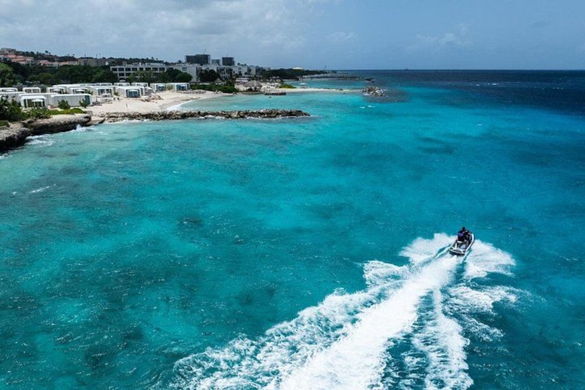 Een jetski op turquoise water, een strand en gebouwen op de achtergrond. 3