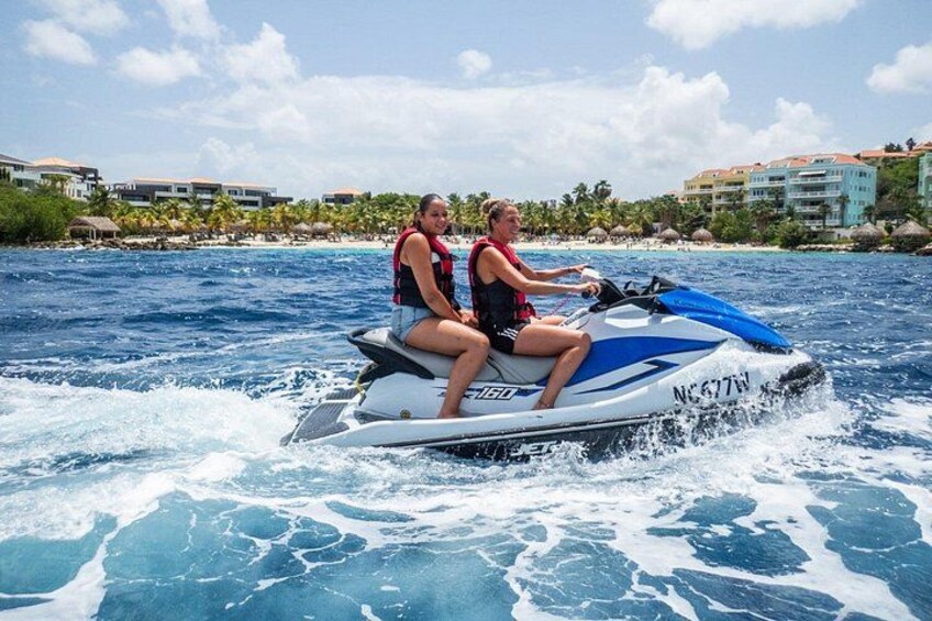 Twee personen op een jetski op het water, met gebouwen op de achtergrond. 1