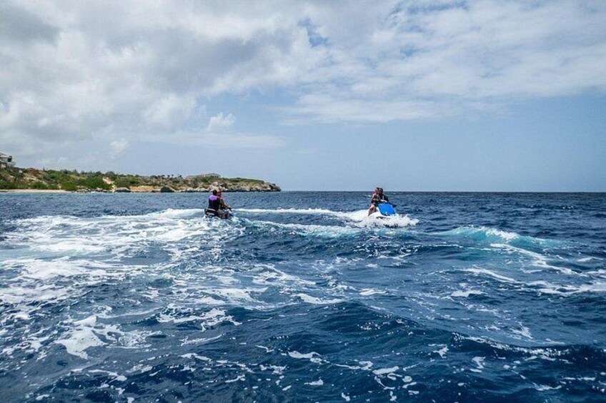 Twee mensen op een surfplank in de oceaan, met een kustlijn op de achtergrond. 2