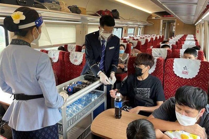 En tågvagn med passagerare som sitter på sina platser, en tågvärd som serverar drycker och en vagn med mat och dryck. 5