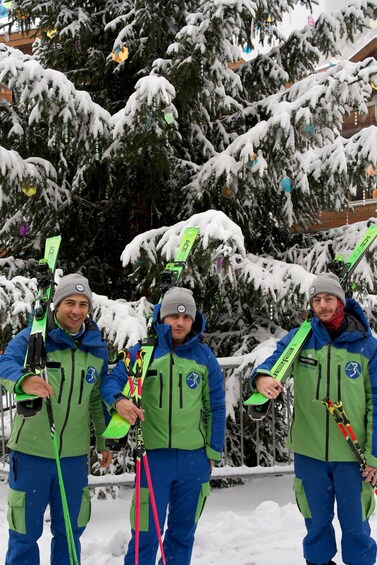 Three people in blue and green ski gear holding skis in front of a snow-covered tree. 2