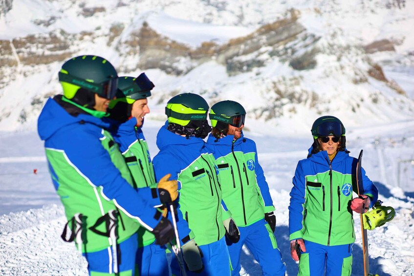 A group of skiers in blue and green jackets with helmets, standing on a snowy slope. 1