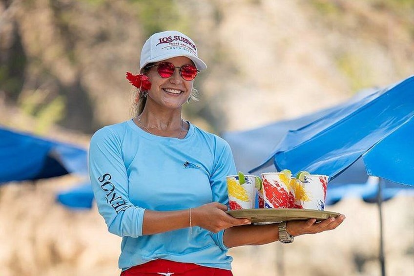 A person holding a tray with colorful drinks and a plate of food, wearing a light blue shirt and a cap with text. 3