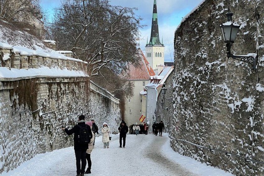 En snedækket gade med folk, en kirke med et grønt tårn og stenvægge. 5