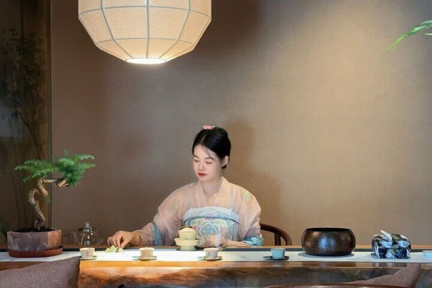 Eine Person in traditioneller Kleidung sitzt an einem Tisch mit einem Teekessel, Tassen und einem Bonsai-Baum. 2