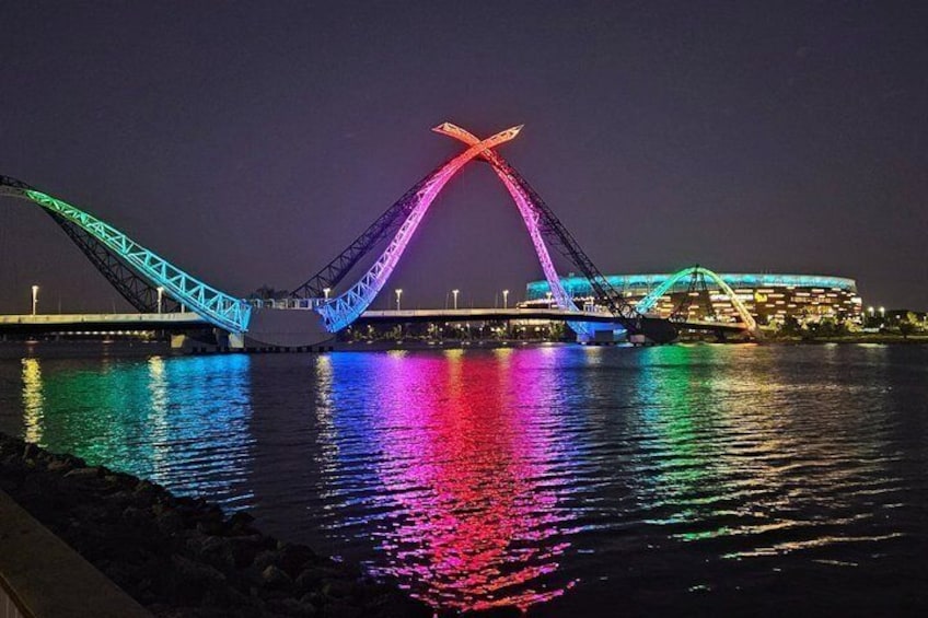 A brightly lit bridge with a distinctive X-shaped structure, reflecting on the water. 1