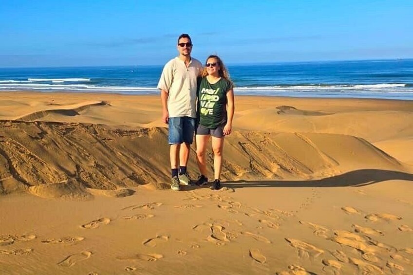 Two people standing on a sandy beach with footprints, ocean waves, and clear skies in the background. 2