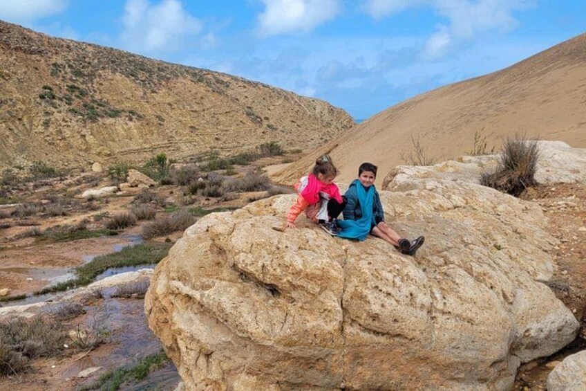 Two people sitting on a large rock in a dry, hilly landscape with sparse vegetation. 5
