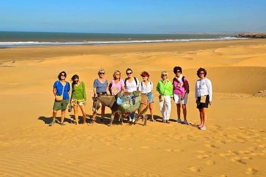 A group of people standing on a sandy beach with a camel in the center. 4