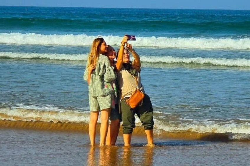 A beach with waves, people standing in shallow water, and a person taking a photo. 3