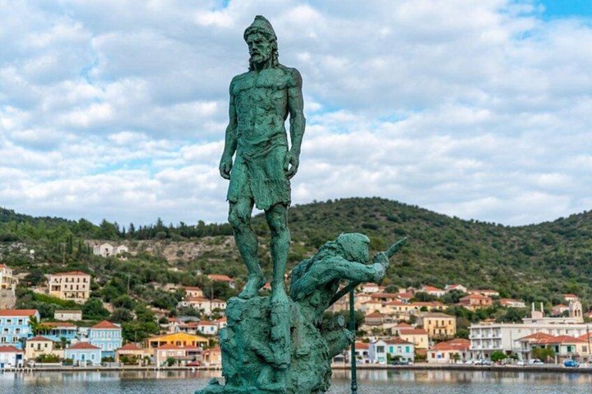 Une statue représentant un homme debout sur un rocher, avec une autre figure, dans une pose dynamique, en contrebas. L’ensemble est placé devant un paysage représentant une ville côtière, avec ses bâtiments et ses collines. 1