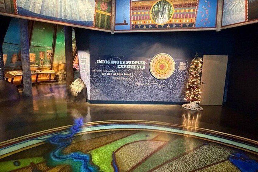 En udstilling med titlen 'INDIGENOUS PEOPLES EXPERIENCE', et cirkulært emblem, et juletræ og et spejlende gulv. 2