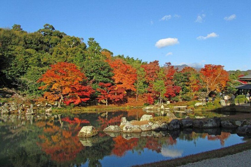 A serene landscape with a pond reflecting trees with autumn foliage, a stone bridge, and a traditional building in the background. 5