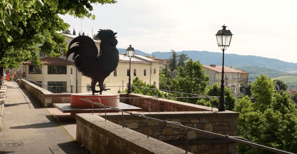 A rooster statue on a stone platform with a railing, overlooking a town with buildings and hills in the background. 2