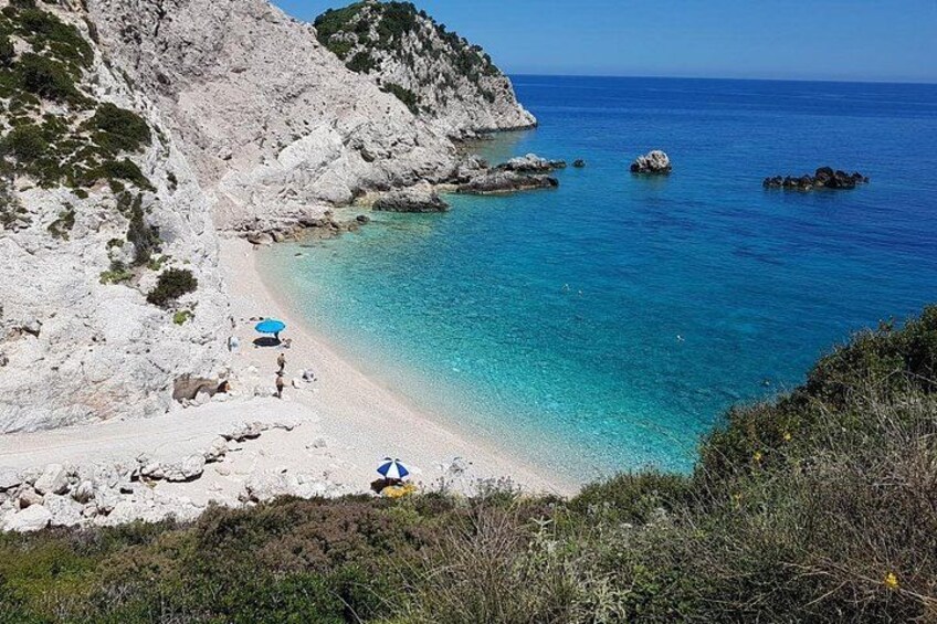 A coastal landscape with a beach, clear blue water, rocky cliffs, and sparse vegetation. 3